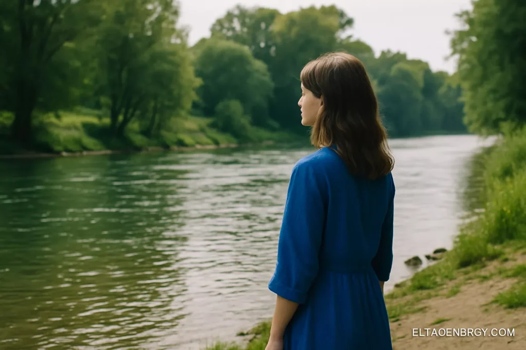 A woman standing near a riverbank, dressed in blue, with peaceful energy.